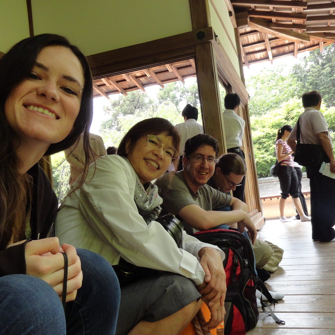 Four people sit in a row on the outdoor porch of a wooden house. Three of them are looking at the camera and smiling, the last person in the row is looking down. There are several other people in the background of the photo, facing away from the camera