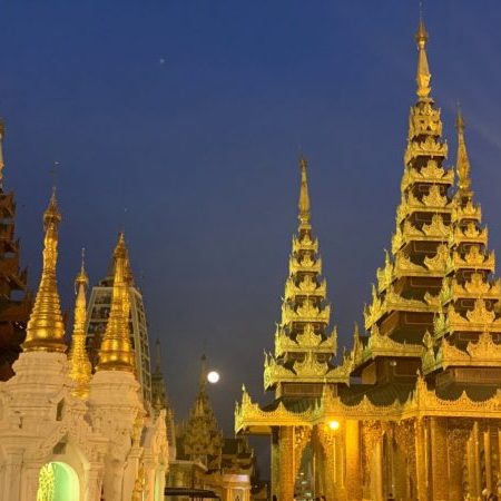 Tall buildings with point spires in Myanmar. The sky is dark blue and looks with dusk with the moon visible in the background. The bulindings are lit up and are a soft yellow color